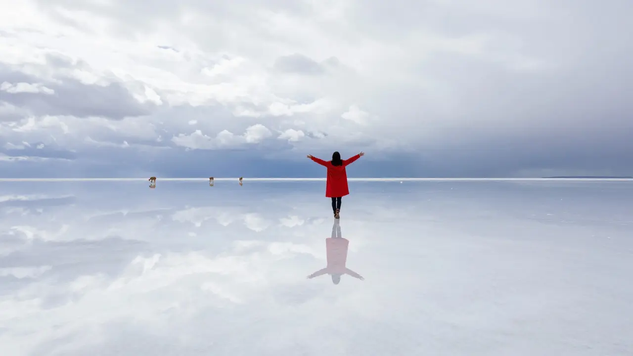 A person standing on the mirror-like salt flat of Salar de Uyuni, reflecting a cloud-filled sky with distant llamas.